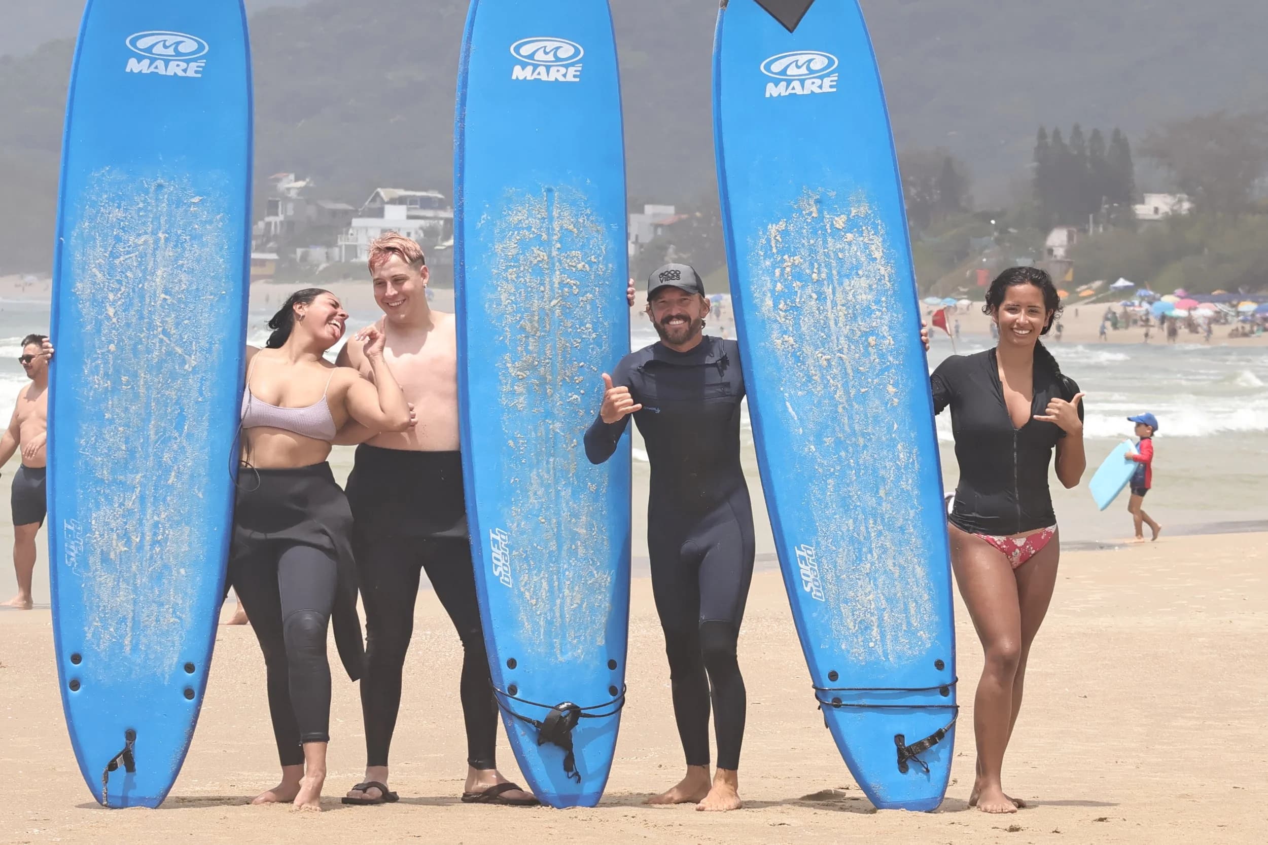 Quatro amigos com pranchas de surf azuis na areia da praia, fazendo o sinal de shaka
